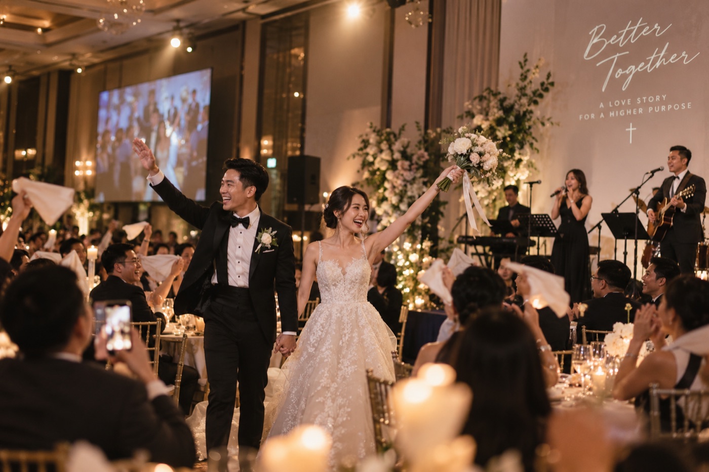 Newlyweds marching into their wedding reception in Singapore as the live band plays on stage behind them and guests wave napkins.